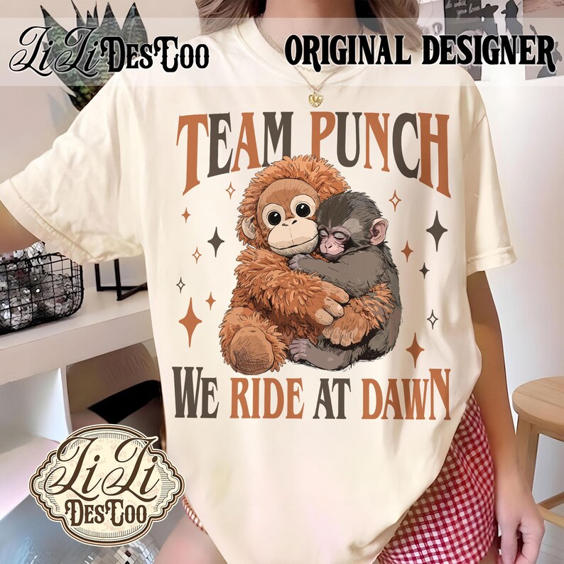 May include: Cream-colored t-shirt featuring a graphic of two monkeys hugging, with the text "TEAM PUNCH" and "WE RIDE AT DAWN" in brown. The shirt also has the text "ORIGINAL DESIGNER" and the brand logo "Li Li DesToo".