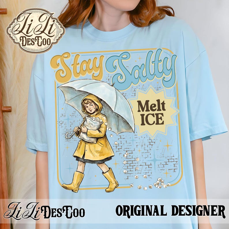 Pode incluir: Camiseta azul clara com um gr&aacute;fico de uma menina de capa de chuva e botas amarelas sob um guarda-chuva. O texto "Stay Salty Melt ICE" est&aacute; em estilo retr&ocirc;. A camisa tamb&eacute;m apresenta o logotipo "Ji Ji DesToo".