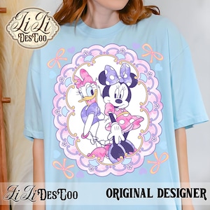 May include: Light blue t-shirt featuring a framed illustration of Minnie Mouse and Daisy Duck. The design includes pink, purple, and yellow accents. The shirt has the text "ORIGINAL DESIGNER" and a logo.