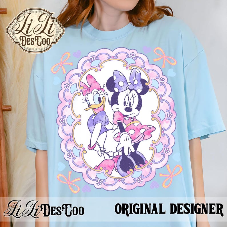 May include: Light blue t-shirt featuring a framed illustration of Minnie Mouse and Daisy Duck. The design includes pink, purple, and yellow accents. The shirt has the text "ORIGINAL DESIGNER" and a logo.