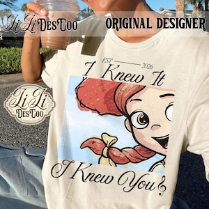 K&ouml;nnte beinhalten: Ein cremefarbenes T-Shirt mit einer Grafik einer Zeichentrickfigur mit roten Haaren im Pferdeschwanz. Der Text lautet "EST 2026", "I Knew It" und "I Knew You" mit einer Musiknote. Das Design ist auf der Vorderseite des Shirts zentriert.