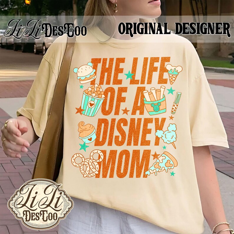 May include: Cream-colored t-shirt with the text "THE LIFE OF A DISNEY MOM" in orange, featuring food-themed graphics. The shirt includes images of a burger, popcorn, ice cream, and pizza. The brand name "Li Li DesCoo" is also visible.