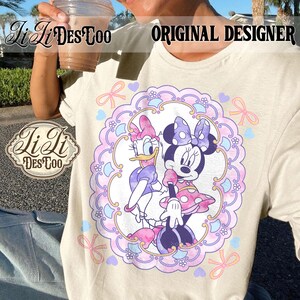 May include: Cream-colored t-shirt featuring a circular design with Minnie Mouse and Daisy Duck. The design includes a pink and purple scalloped border with bows and hearts. The words "ORIGINAL DESIGNER" are printed on the shirt.