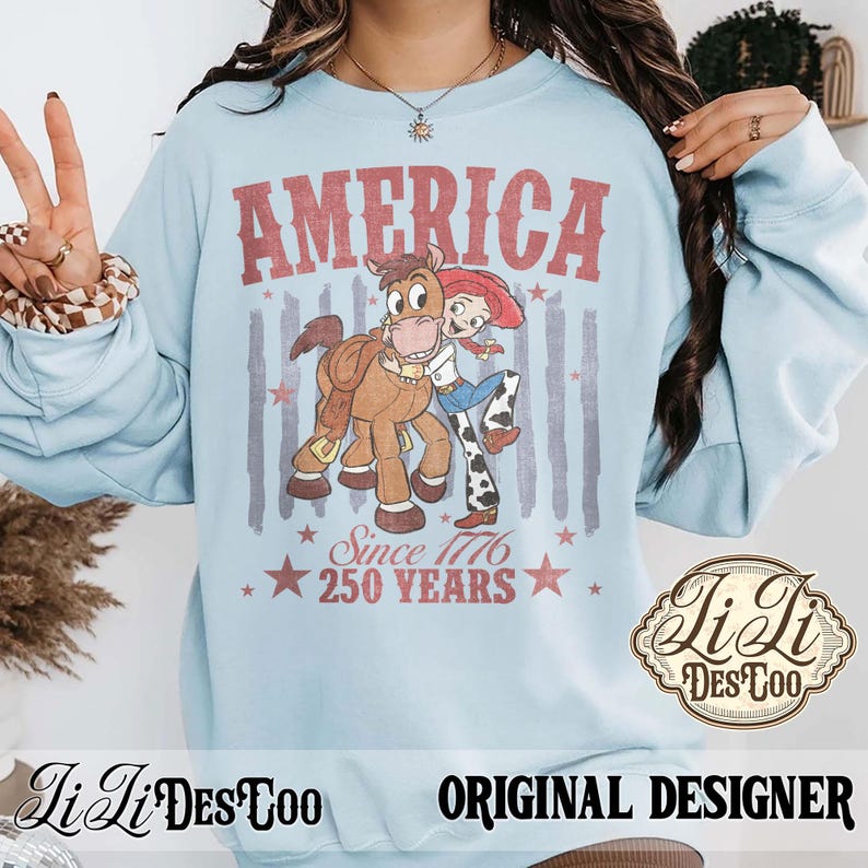 May include: A light blue crewneck sweatshirt featuring a graphic of Jessie the cowgirl hugging Bullseye the horse. The text reads AMERICA, Since 1776, and 250 YEARS in red. The design includes faded red and blue vertical stripes and stars.