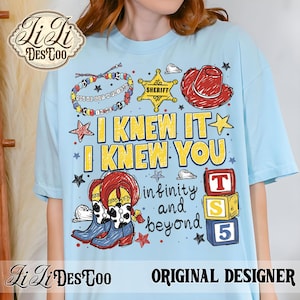 Op de afbeelding: Een lichtblauw t-shirt met een grafische print van een kralenarmband, een gele sheriffster, een rode cowboyhoed en laarzen met koeienvlekken. De tekst luidt I KNEW IT I KNEW YOU, infinity and beyond, en TS 5 op gestapelde blokken.