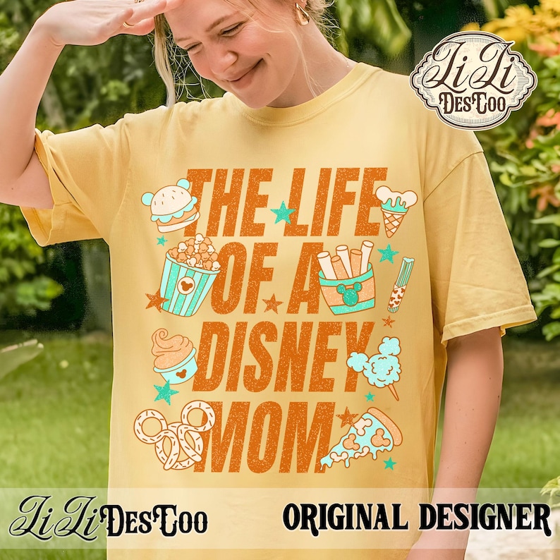 May include: Mustard yellow t-shirt with the text "THE LIFE OF A DISNEY MOM" in orange. The shirt features cartoon illustrations of a hamburger, popcorn, ice cream, fries, a candy stick, cotton candy, a pretzel, and a pizza.