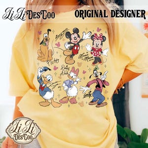 Puede incluir: Camiseta amarilla con un gr&aacute;fico colorido de personajes de Disney, incluyendo Mickey Mouse, Minnie Mouse, Pluto, Donald Duck, Daisy Duck y Goofy. La camiseta tiene el texto "Li Li Des Too" y "Original Designer".