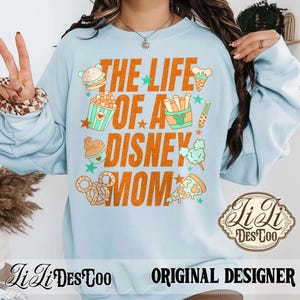 May include: Light blue sweatshirt with the text "THE LIFE OF A DISNEY MOM" in orange. The design includes illustrations of food items like a burger, popcorn, ice cream, fries, cotton candy, pizza, and pretzels. The sweatshirt also has the text "Li Li DesToo" and "ORIGINAL DESIGNER".