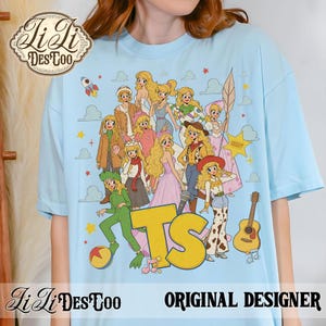 May include: A light blue t-shirt featuring a graphic print of multiple blonde characters in various outfits, including a green dinosaur costume, a cowgirl ensemble, and a space suit. The design includes a large yellow TS logo, a guitar, and a ball.