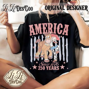 May include: A black t-shirt featuring a graphic of a cartoon cowgirl hugging a horse. The text reads AMERICA, Since 1776, and 250 YEARS in pink lettering. The design includes vertical distressed stripes and star accents, creating a patriotic theme.