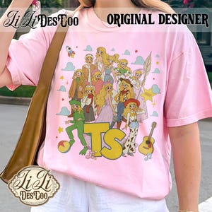 May include: A light pink t-shirt featuring a graphic print of multiple blonde characters in various outfits, including a green suit, a cowboy hat, and a crown. The design includes the yellow letters TS, a guitar, a ball, and clouds.