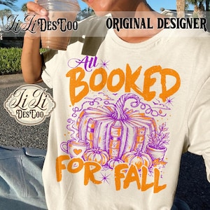 May include: A cream-colored t-shirt featuring a graphic print with the text "All BOOKED FOR FALL" in orange. The design includes a pumpkin made of stacked books, a steaming mug with a heart, small pumpkins, and a potted plant in purple and orange.
