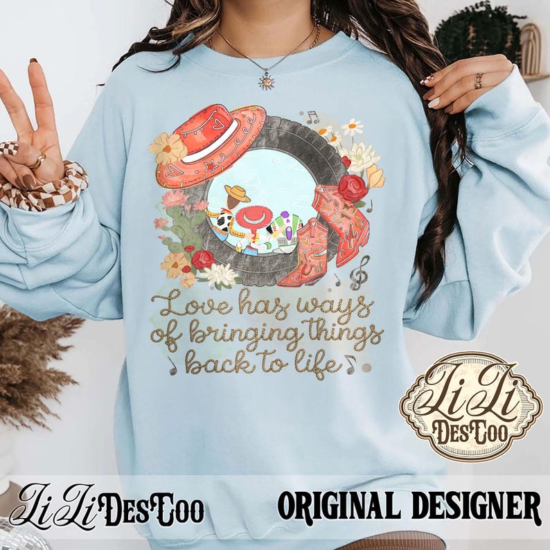 Puede incluir: Una sudadera de cuello redondo azul claro con un estampado gr&aacute;fico de un marco de neum&aacute;tico que contiene personajes de dibujos animados, un sombrero rojo y botas vaqueras, rodeado de flores. El texto dice, Love has ways of bringing things back to life.
