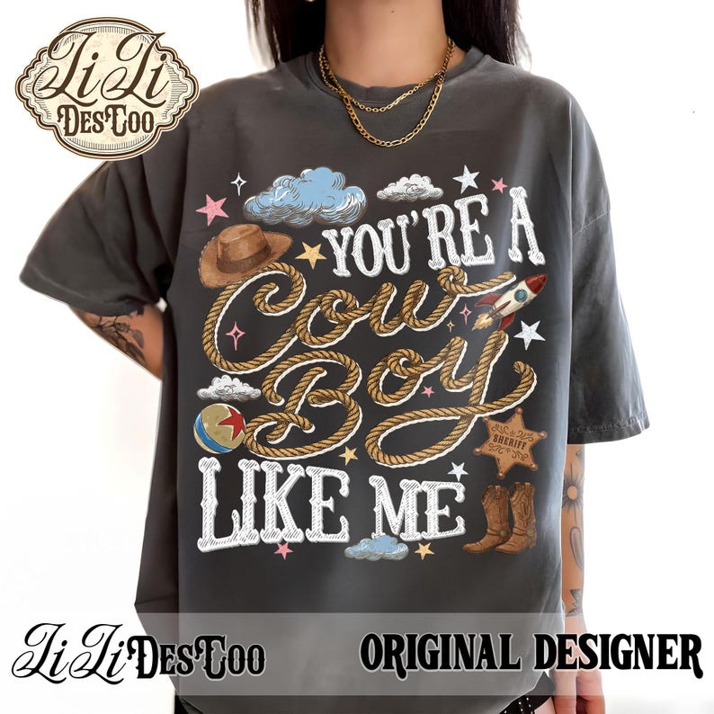 May include: A charcoal gray t-shirt featuring a graphic print with the text "YOU'RE A COWBOY LIKE ME" in white lettering. The design includes a brown cowboy hat, a rope, a rocket, a sheriff star, a colorful ball, and blue clouds with small stars.