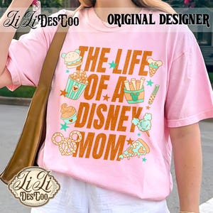 May include: Pink t-shirt with the text "THE LIFE OF A DISNEY MOM" in orange. The shirt features cartoon illustrations of food items like a burger, popcorn, ice cream, and pizza. The words "ORIGINAL DESIGNER" are printed at the top.