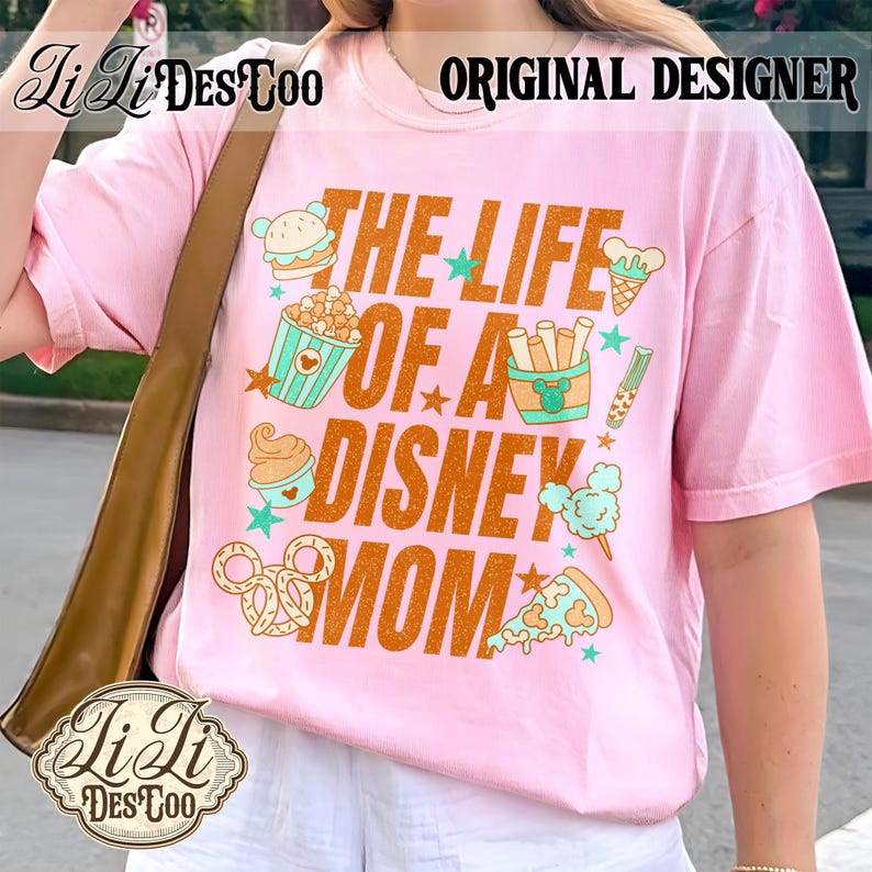 May include: Pink t-shirt with the text "THE LIFE OF A DISNEY MOM" in orange. The shirt features cartoon illustrations of food items like a burger, popcorn, ice cream, and pizza. The words "ORIGINAL DESIGNER" are printed at the top.