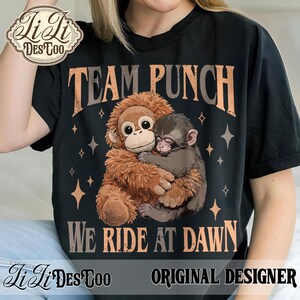 May include: Black t-shirt featuring a graphic of two monkeys hugging, with the text "TEAM PUNCH" above and "WE RIDE AT DAWN" below. The design includes star accents and the words "ORIGINAL DESIGNER".