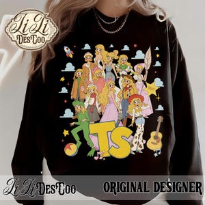 May include: A black long-sleeve sweatshirt featuring a graphic print of multiple blonde characters in various outfits, including a green alien costume, a cowgirl outfit, and a space ranger suit. The text TS is printed in large yellow letters.