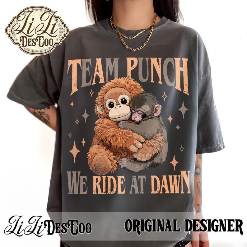 May include: Dark gray t-shirt featuring a graphic of two monkeys hugging, with the text "TEAM PUNCH" and "WE RIDE AT DAWN" in orange and gray. The shirt also has the text "Li Li Des Too" and "ORIGINAL DESIGNER".
