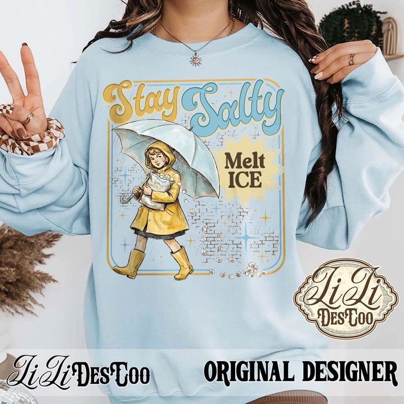 Pode incluir: Camisola azul clara com um gr&aacute;fico que diz "Stay Salty Melt Ice". O gr&aacute;fico apresenta uma crian&ccedil;a com capa de chuva e botas amarelas segurando um guarda-chuva. O design &eacute; emoldurado com uma borda dourada e inclui o logotipo do designer.