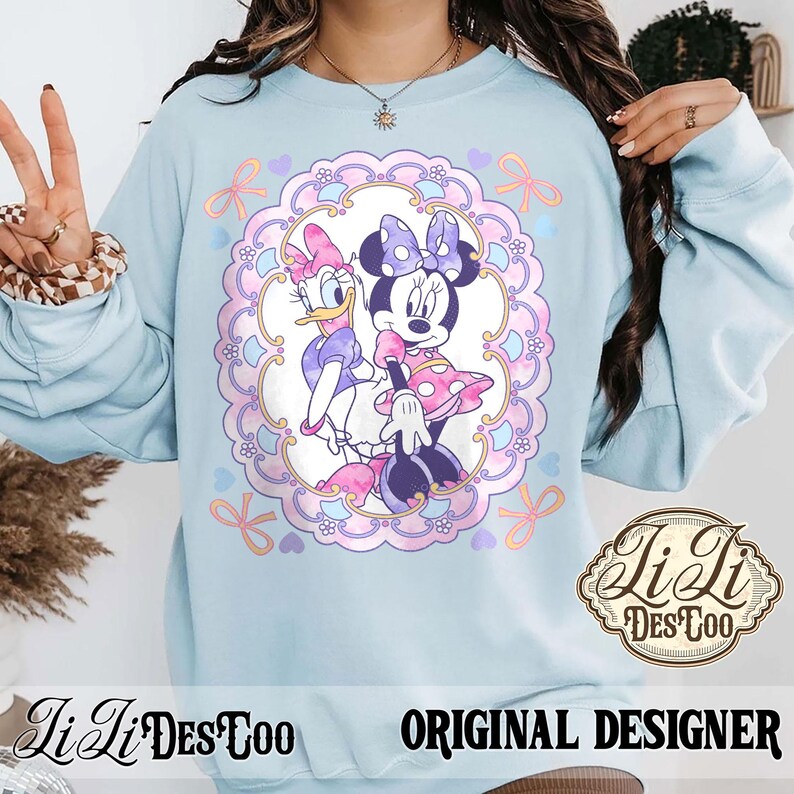 May include: Light blue sweatshirt featuring a framed illustration of Minnie Mouse and Daisy Duck. The illustration is surrounded by a pink and purple floral design with bows and hearts. The sweatshirt has the text "LiLi DesToo" and "ORIGINAL DESIGNER".