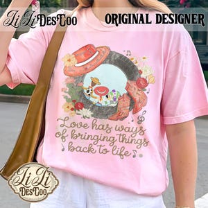 Puede incluir: Una camiseta rosa claro con un estampado gr&aacute;fico de un marco de neum&aacute;tico que contiene personajes de dibujos animados, un sombrero de vaquero rojo y botas vaqueras naranjas rodeadas de flores. El texto dice, Love has ways of bringing things back to life.