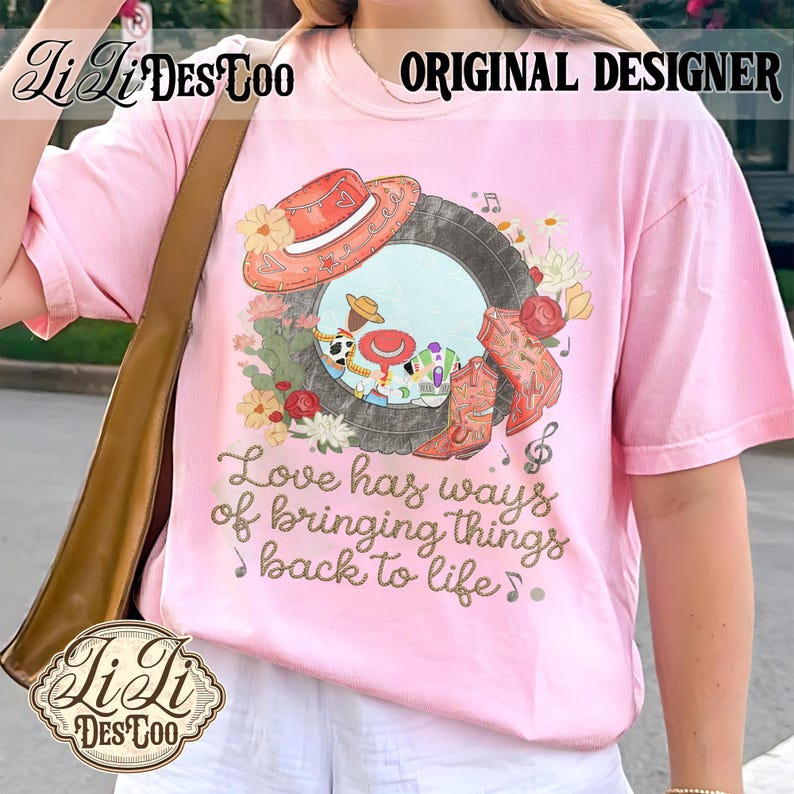 Puede incluir: Una camiseta rosa claro con un estampado gr&aacute;fico de un marco de neum&aacute;tico que contiene personajes de dibujos animados, un sombrero de vaquero rojo y botas vaqueras naranjas rodeadas de flores. El texto dice, Love has ways of bringing things back to life.