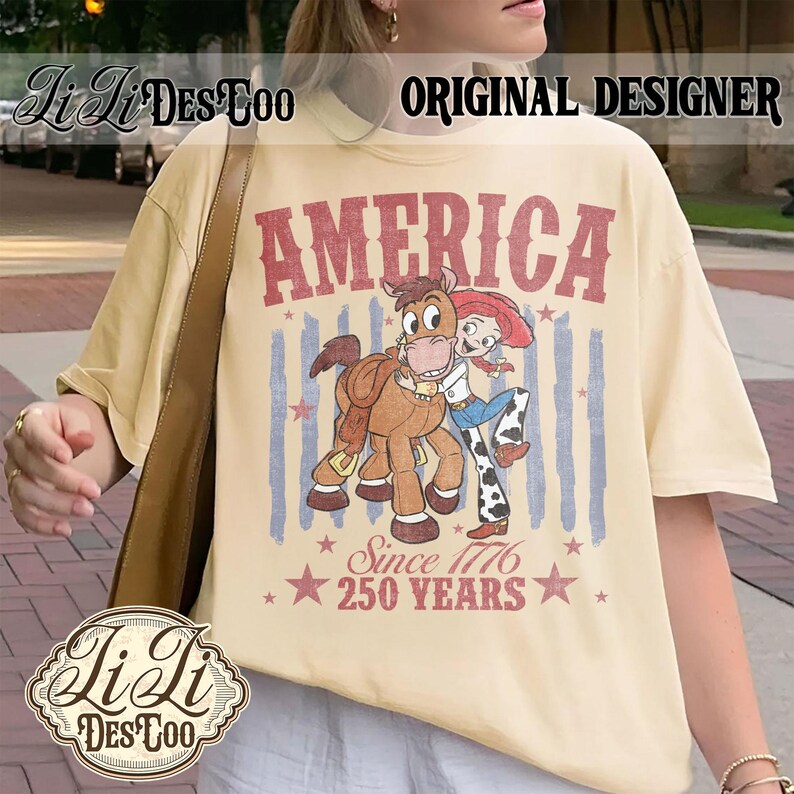 May include: A cream-colored t-shirt featuring a graphic of a cowgirl character hugging a brown horse. The text reads AMERICA, Since 1776, and 250 YEARS, accompanied by red and blue vertical stripes and stars. The design has a distressed, vintage-inspired look.