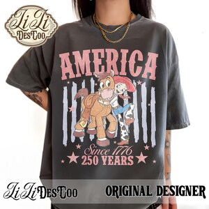 May include: A charcoal gray t-shirt featuring a graphic of a cowgirl and a horse. The text reads AMERICA, Since 1776, and 250 YEARS. The design includes faded vertical stripes and stars in a muted pink tone, with a distressed, vintage-inspired aesthetic.