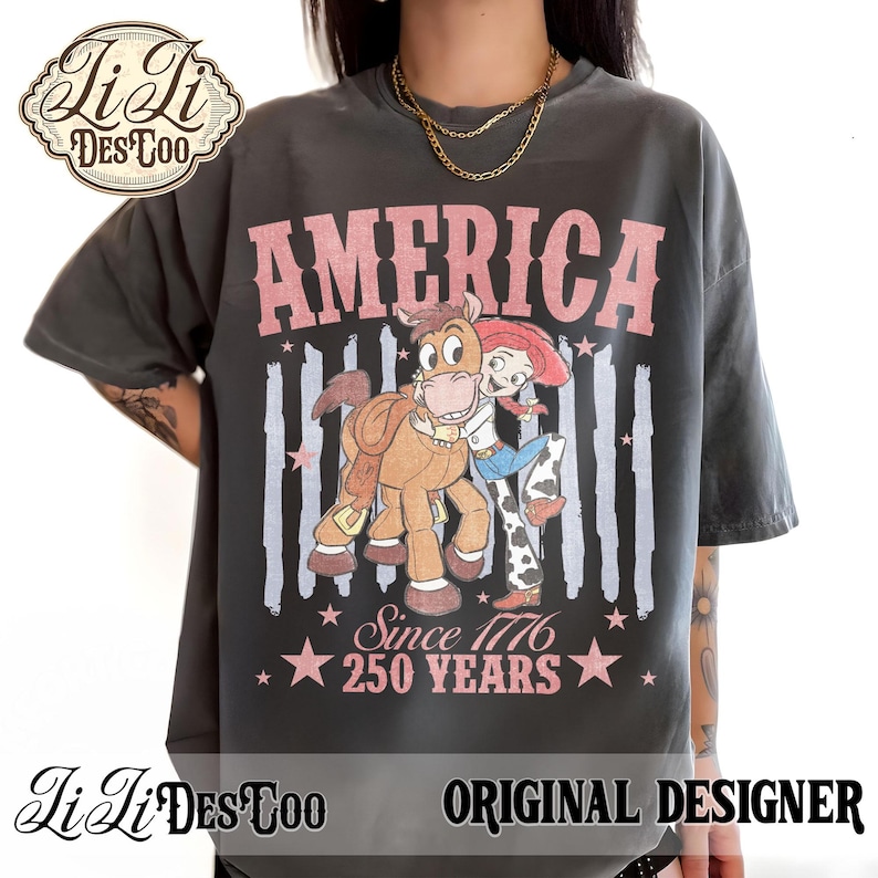 May include: A charcoal gray t-shirt featuring a graphic of a cowgirl and a horse. The text reads AMERICA, Since 1776, and 250 YEARS. The design includes faded vertical stripes and stars in a muted pink tone, with a distressed, vintage-inspired aesthetic.