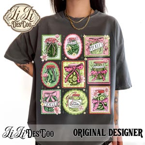May include: Dark gray t-shirt featuring a grid of pickle jar illustrations with pink bows and floral accents. The word "PICKLES" is displayed on some jars. The shirt also has the text "LiLi DES COO ORIGINAL DESIGNER".