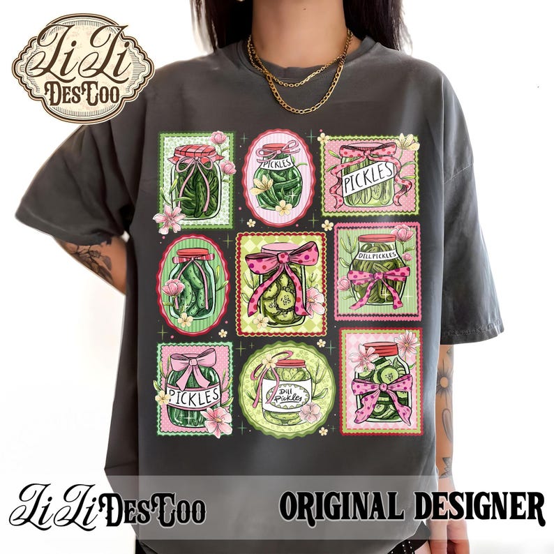 May include: Dark gray t-shirt featuring a grid of pickle jar illustrations with pink bows and floral accents. The word "PICKLES" is displayed on some jars. The shirt also has the text "LiLi DES COO ORIGINAL DESIGNER".
