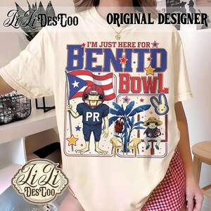 May include: Cream-colored t-shirt with a graphic featuring a frog in a football uniform holding a Puerto Rican flag. Text reads "I'M JUST HERE FOR BENITO BOWL" in blue and red. The shirt has the "Li Ji Des Too" logo.