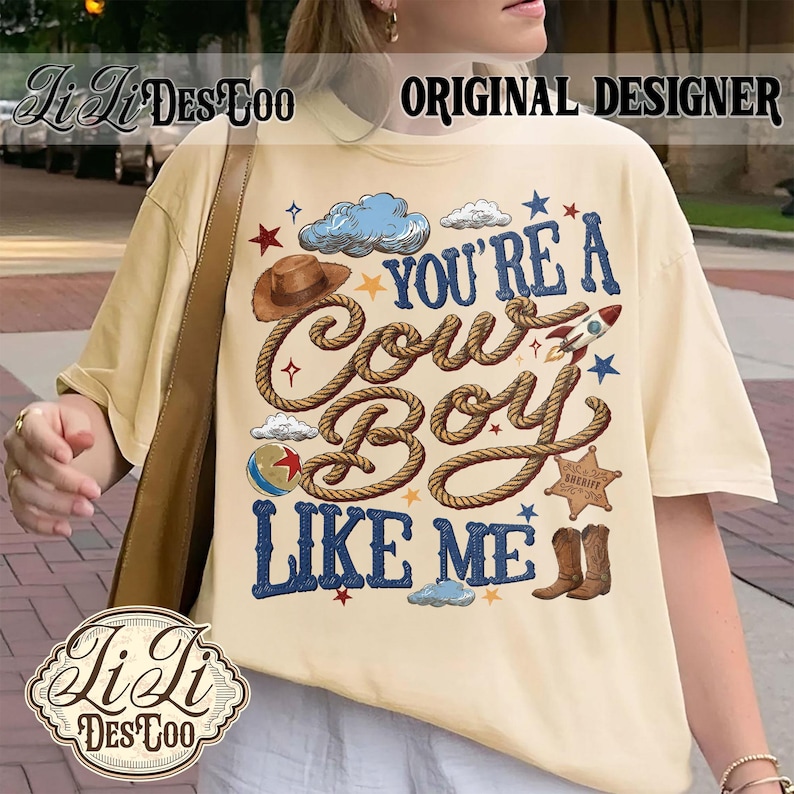 May include: A cream-colored short-sleeve t-shirt featuring a graphic print with the text "YOU'RE A COWBOY LIKE ME" in blue. The design includes a brown cowboy hat, a rope, a rocket, a sheriff star, a ball, and blue clouds on a light background.