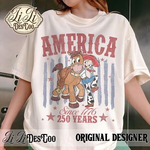 May include: A cream-colored t-shirt featuring a graphic of a cartoon cowgirl hugging a horse. The text reads AMERICA, Since 1776, and 250 YEARS in red. The design includes faded blue vertical stripes and red stars, with a Li Li DES TOO logo in the corner.