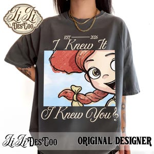 K&ouml;nnte beinhalten: Ein anthrazitfarbenes T-Shirt mit einem Grafikdruck einer Zeichentrickfigur mit roten Haaren und einem Zopf. Der Text lautet EST 2026, I Knew It und I Knew You. Das Design enth&auml;lt einen hellblauen Himmelshintergrund. Das Shirt hat einen Rundhalsausschnitt.