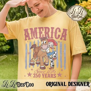 May include: A mustard yellow short-sleeve t-shirt featuring a graphic print with the text AMERICA, SINCE 1776, and 250 YEARS. The design includes a cartoon cowgirl hugging a brown horse, set against faded blue vertical stripes and red stars.