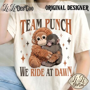 May include: Cream-colored t-shirt with the text "TEAM PUNCH WE RIDE AT DAWN" in brown and black. The shirt features a graphic of two hugging monkeys. The design includes star shapes. "Li Li Des Too" and "Original Designer" are also printed on the shirt.