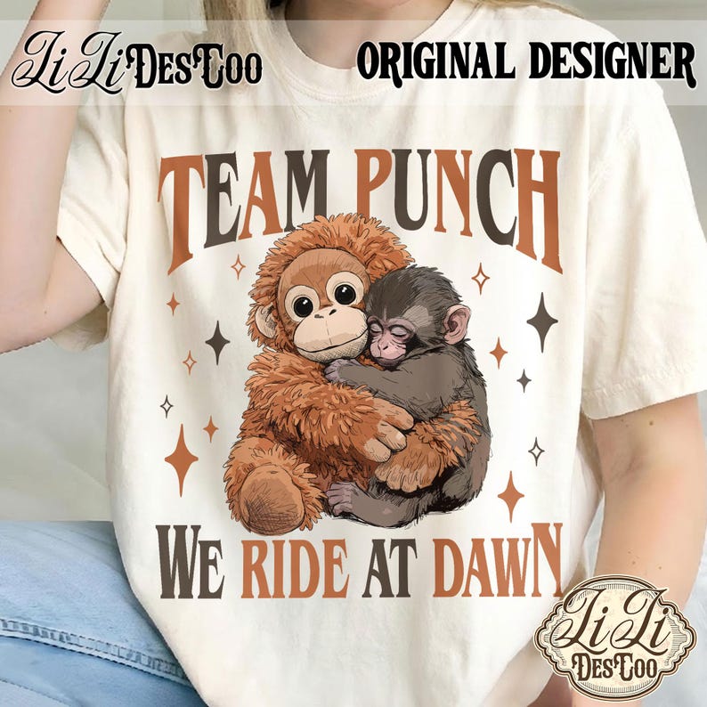 May include: Cream-colored t-shirt with the text "TEAM PUNCH WE RIDE AT DAWN" in brown and black. The shirt features a graphic of two hugging monkeys. The design includes star shapes. "Li Li Des Too" and "Original Designer" are also printed on the shirt.