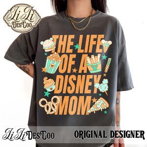 May include: Dark gray t-shirt with the text "THE LIFE OF A DISNEY MOM" in orange. The shirt features illustrations of Disney-themed snacks like popcorn, ice cream, and pretzels. The shirt has the "JiLi DesToo" logo and "ORIGINAL DESIGNER" text.