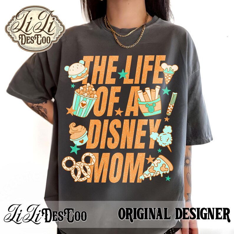 May include: Dark gray t-shirt with the text "THE LIFE OF A DISNEY MOM" in orange. The shirt features illustrations of Disney-themed snacks like popcorn, ice cream, and pretzels. The shirt has the "JiLi DesToo" logo and "ORIGINAL DESIGNER" text.