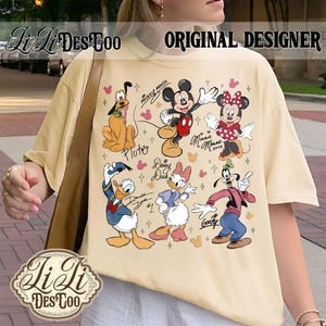 Puede incluir: Camiseta color crema con un gr&aacute;fico colorido de personajes de Disney, incluyendo Mickey Mouse, Minnie Mouse, Pluto, Donald Duck, Daisy Duck y Goofy. La camiseta tiene el texto "Li Li Des Too" y "Original Designer".