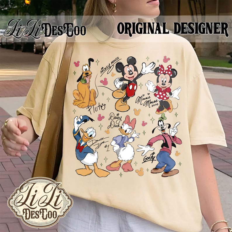 Puede incluir: Camiseta color crema con un gr&aacute;fico colorido de personajes de Disney, incluyendo Mickey Mouse, Minnie Mouse, Pluto, Donald Duck, Daisy Duck y Goofy. La camiseta tiene el texto "Li Li Des Too" y "Original Designer".
