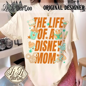 May include: Cream-colored t-shirt with the text "THE LIFE OF A DISNEY MOM" in orange, featuring cartoon food icons like a burger, popcorn, ice cream, and pretzels. The shirt also has the text "ORIGINAL DESIGNER".