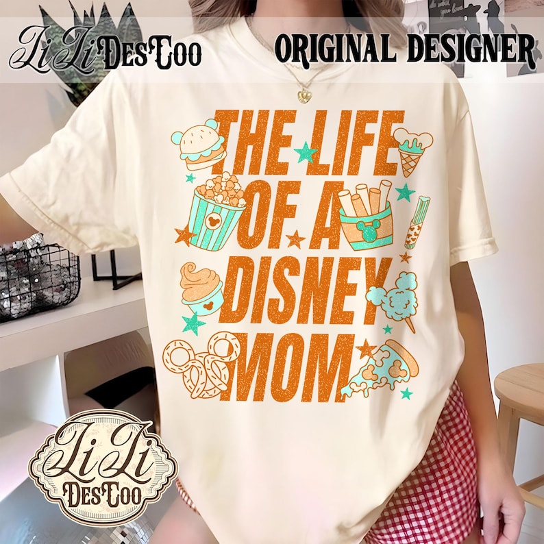 May include: Cream-colored t-shirt with the text "THE LIFE OF A DISNEY MOM" in orange, featuring cartoon food icons like a burger, popcorn, ice cream, and pretzels. The shirt also has the text "ORIGINAL DESIGNER".
