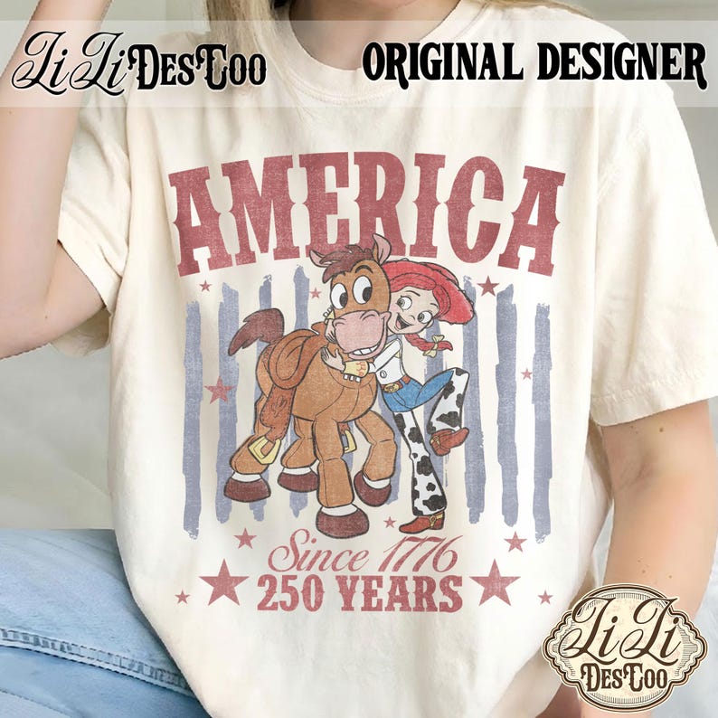 May include: A cream-colored short-sleeve t-shirt featuring a graphic of a cowgirl hugging a brown horse. The text reads AMERICA, Since 1776, 250 YEARS. The design includes faded blue vertical stripes and red stars, with the brand name LiLi DES TOO.