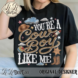 May include: A black t-shirt featuring the text "YOU'RE A COWBOY LIKE ME" written in white rope-style lettering. The design includes a brown cowboy hat, a rocket, a sheriff star badge, a ball, clouds, and stars in various colors.