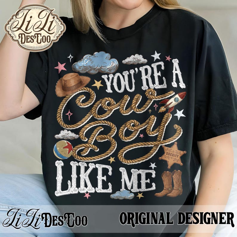 May include: A black t-shirt featuring the text "YOU'RE A COWBOY LIKE ME" written in white rope-style lettering. The design includes a brown cowboy hat, a rocket, a sheriff star badge, a ball, clouds, and stars in various colors.