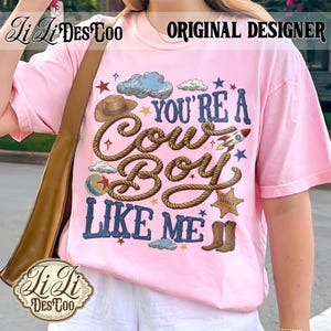 May include: A light pink t-shirt featuring a graphic print with the text "YOU'RE A COWBOY LIKE ME" in blue lettering. The design includes a brown cowboy hat, a rope, a rocket, a sheriff star badge, a ball, and brown cowboy boots with blue clouds.