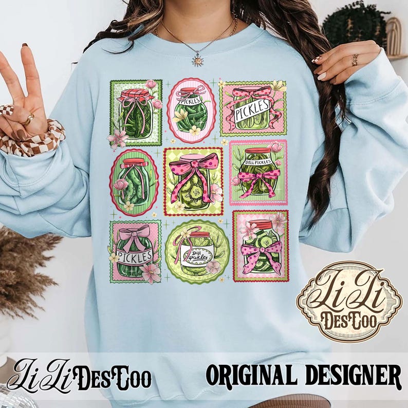 May include: Light blue sweatshirt featuring a grid of pickle jar illustrations with pink bows and floral accents. The word "PICKLES" is displayed on several jars. The design includes the text "Li Li Des Too" and "ORIGINAL DESIGNER".
